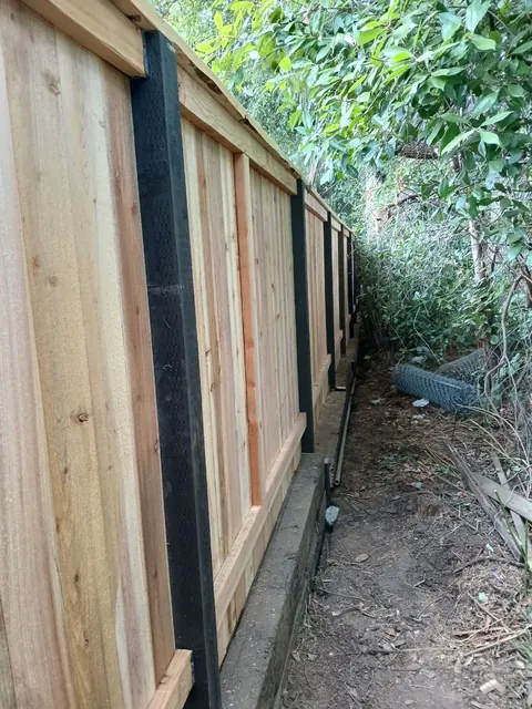 Vertical wood boards are supported by horizontal rails and dark posts along a narrow planted boundary.